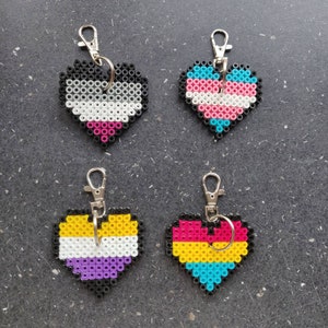 Könnte beinhalten: Vier herzförmige Schlüsselanhänger aus bunten Perlen. Jeder Schlüsselanhänger zeigt eine andere Pride-Flagge: grau, weiß, rosa und schwarz; hellblau, rosa, weiß und dunkelblau; gelb, weiß, lila und schwarz; und gelb, hellblau, rosa und dunkelblau.