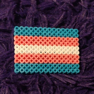 May include: A small, rectangular trans pride flag made of blue, pink, and white beads. The flag is made of small, round beads that are strung together to form the flag.