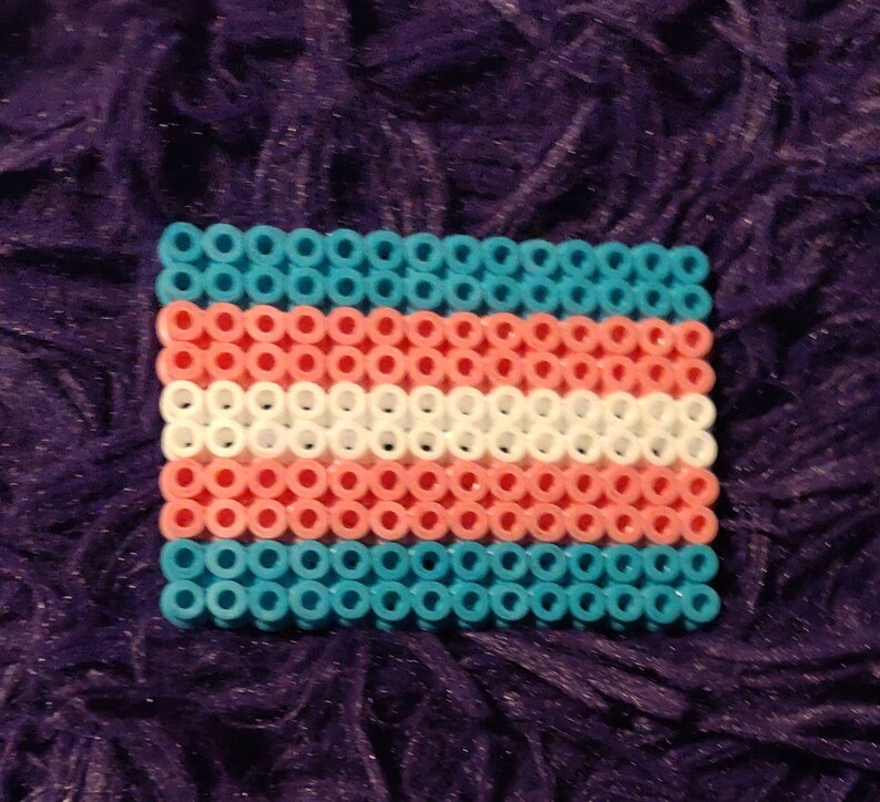 May include: A small, rectangular trans pride flag made of blue, pink, and white beads. The flag is made of small, round beads that are strung together to form the flag.