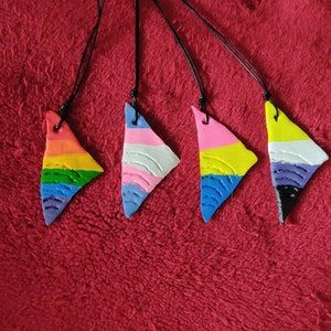 Könnte beinhalten: Vier dreieckige Anhänger mit verschiedenen Pride-Flaggen-Designs. Die Anhänger sind aus Ton gefertigt und haben eine schwarze Schnur daran befestigt. Die Flaggen sind Regenbogen, Transgender, Bisexuell und Non-Binary.