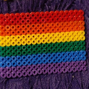 May include: A rainbow pride flag made of colorful round beads. The flag is made of six rows of beads, with each row representing a different color of the rainbow: red, orange, yellow, green, blue, and purple.