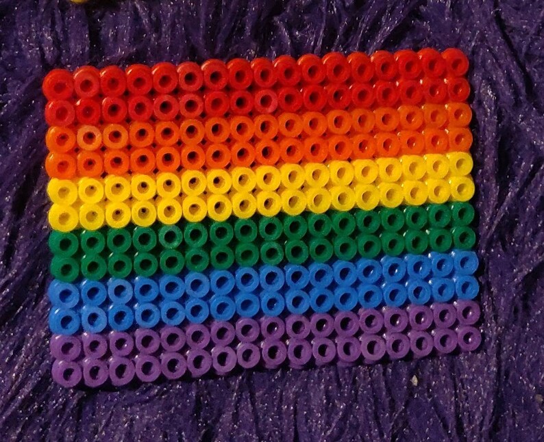 May include: A rainbow pride flag made of colorful round beads. The flag is made of six rows of beads, with each row representing a different color of the rainbow: red, orange, yellow, green, blue, and purple.