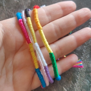 Könnte beinhalten: Eine Sammlung farbenfroher Freundschaftsarmbänder, jedes mit leuchtenden Fäden umwickelt. Die Armbänder weisen eine Vielzahl von Farben auf, darunter Rosa, Blau, Gelb, Grün und Lila. Die Enden sind mit kleinen, bunten Perlen versehen.