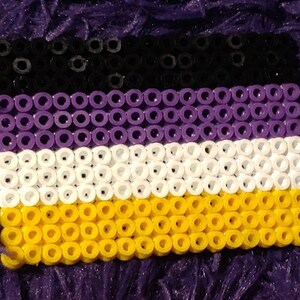 May include: A close-up of a small, rectangular flag made of black, purple, white, and yellow beads. The flag is a representation of the non-binary pride flag.