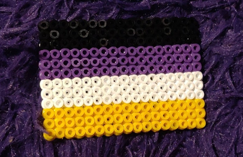 May include: A close-up of a small, rectangular flag made of black, purple, white, and yellow beads. The flag is a representation of the non-binary pride flag.