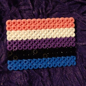 May include: A small, rectangular flag made of colorful beads. The flag has five horizontal stripes: pink, white, purple, black, and blue.
