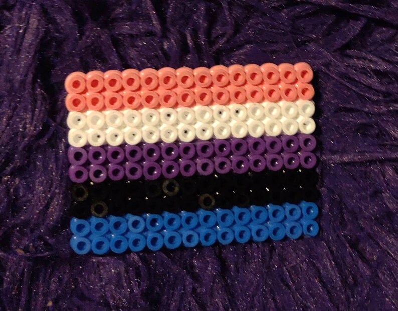 May include: A small, rectangular flag made of colorful beads. The flag has five horizontal stripes: pink, white, purple, black, and blue.