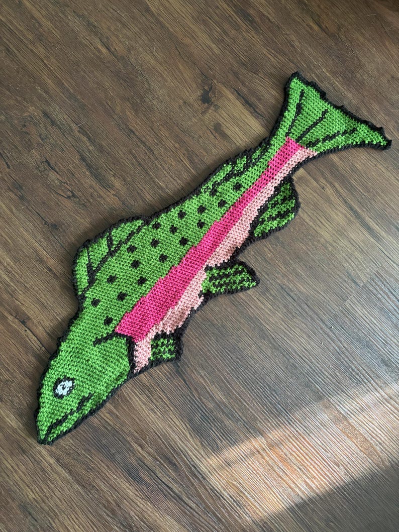 Puede incluir: Una alfombra de pescado de crochet verde y rosa con detalles negros. El pez tiene un ojo blanco y una raya rosa a lo largo de su lado.
