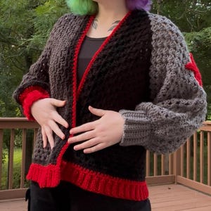 May include: A handmade crochet cardigan in shades of brown, black, and gray, with red accents on the cuffs and trim. The open-front design features a textured stitch pattern, offering a cozy and stylish look. This unique piece is perfect for layering.