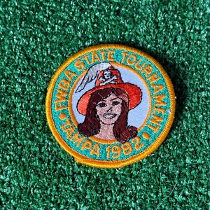 May include: A circular patch with a teal border and gold stitching. The patch features the text "FYBA STATE TOURNAMENT TAMPA 1982" around a central image of a woman wearing a pirate hat.