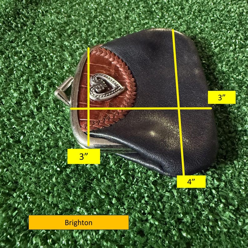 May include: A dark blue leather coin purse with a brown textured leather accent and a silver-tone metal heart emblem. The purse features a metal kiss-lock closure. Yellow lines indicate dimensions of 3 inches by 4 inches. The brand name Brighton is visible.