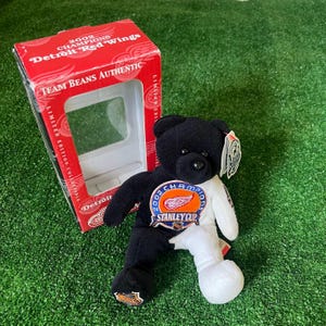May include: A black and white plush bear wearing a Detroit Red Wings jersey with the 2002 Stanley Cup Champions logo. The bear is sitting on a green surface and is in a red and white box with the words "2002 Champions Detroit Red Wings" and "Team Beans Authentic" printed on it.