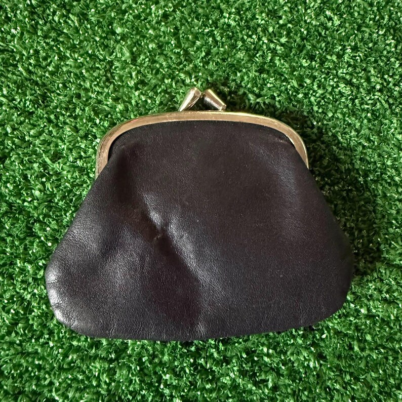 May include: A dark brown leather coin purse featuring a smooth, soft texture and a gold-toned metal kiss-lock clasp closure at the top. The small pouch has a rounded, trapezoidal shape with visible natural grain and subtle creases on the surface.