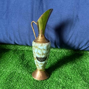 May include: A vintage decorative vase with a brass handle and base. The vase has a light blue and green patterned body and a curved, gold-colored top. The label reads "Old Dutch Delft".