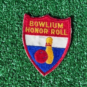 May include: Embroidered patch in a shield shape with the text "BOWLIUM HONOR ROLL". The patch features a red border, white, blue, and red sections, and a bowling pin and ball design. The patch is on a green surface.