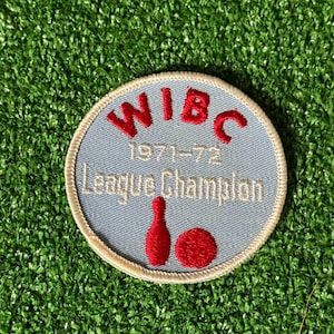 May include: A round patch with a light blue background and a cream-colored border. The patch features the red text "WIBC" above "1971-72 League Champion". Below the text are red bowling pins and a ball.