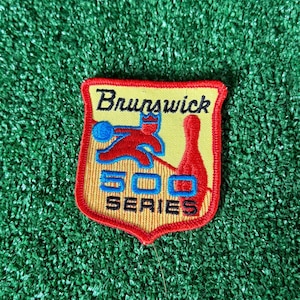 May include: A yellow, red, and black Brunswick 500 Series patch. The patch features a shield shape with the word "Brunswick" at the top. The design includes a bowler, bowling pin, and the text "500 SERIES".