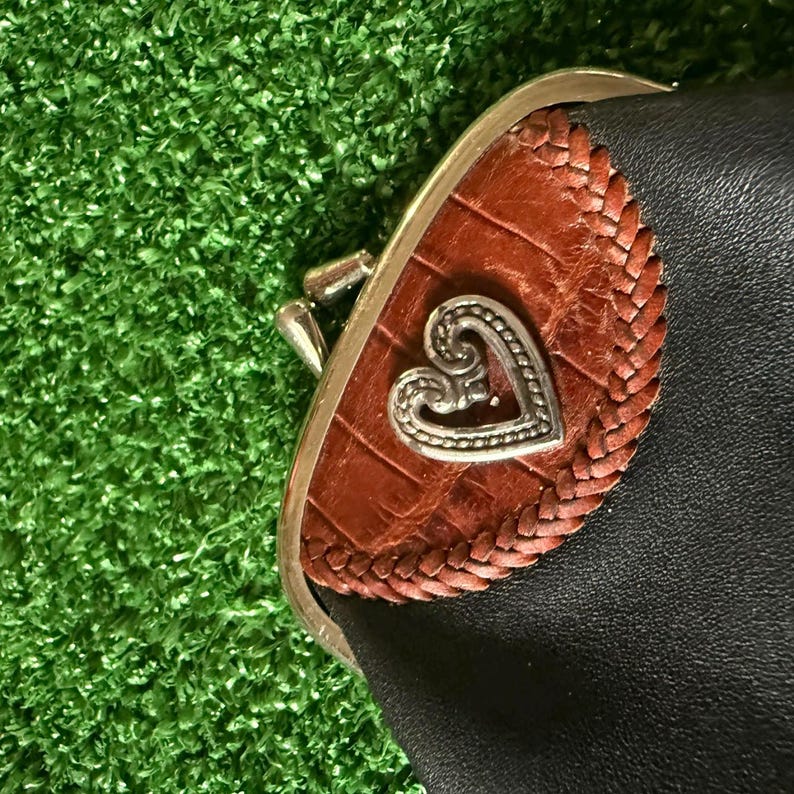 May include: A black coin purse featuring a brown, crocodile-textured leather accent panel with a braided border. A silver-toned metal heart-shaped emblem is centered on the brown panel, and the purse has a silver-toned metal kiss-lock closure.