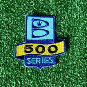May include: Embroidered patch with a blue and yellow design. The patch features the letter "B" with a crown, the number "500", and the words "SERIES". The patch is on a green surface.
