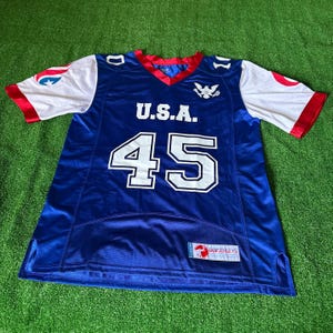 May include: A blue and white football jersey with the text "U.S.A." and the number "45" on the front. The jersey has red and white trim and a small eagle logo on the chest. The jersey is made by "GOF JERSETS".