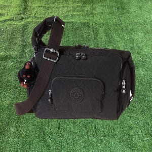 May include: Black shoulder bag with a brown adjustable strap and a small zippered pocket. The bag features a front flap pocket with a circular logo and a small monkey keychain. The bag is made of a durable, water-resistant material.