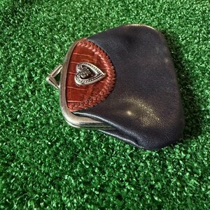 May include: A dark blue leather coin purse featuring a silver-toned metal frame and a clasp closure. The front panel is accented with a brown textured leather insert, a braided leather border, and a central silver-toned heart-shaped emblem.