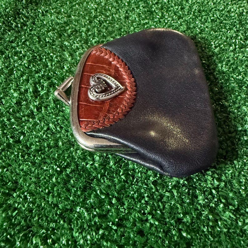 May include: A dark blue leather coin purse featuring a silver-toned metal frame and a clasp closure. The front panel is accented with a brown textured leather insert, a braided leather border, and a central silver-toned heart-shaped emblem.