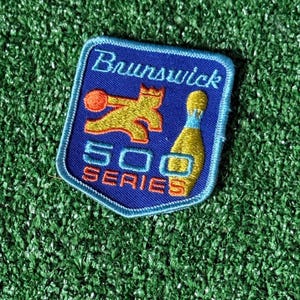 May include: A blue patch with the word "Brunswick" in white script. The patch features a gold bowling pin and a gold figure with a bowling ball. The text "500 SERIES" is in orange.