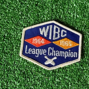 May include: A blue, white, and orange patch with the text "WIBC League Champion" and the years 1964 and 1965. The patch has a white border and a white "X" at the bottom. The background is green.