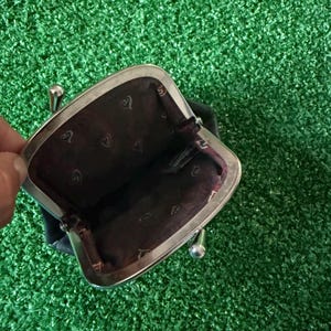 May include: A small black coin purse with a silver-toned metal kiss-lock frame. The interior is lined with dark brown fabric featuring a repeating pattern of small, light-colored heart-shaped icons and a visible brand tag inside the compartment.