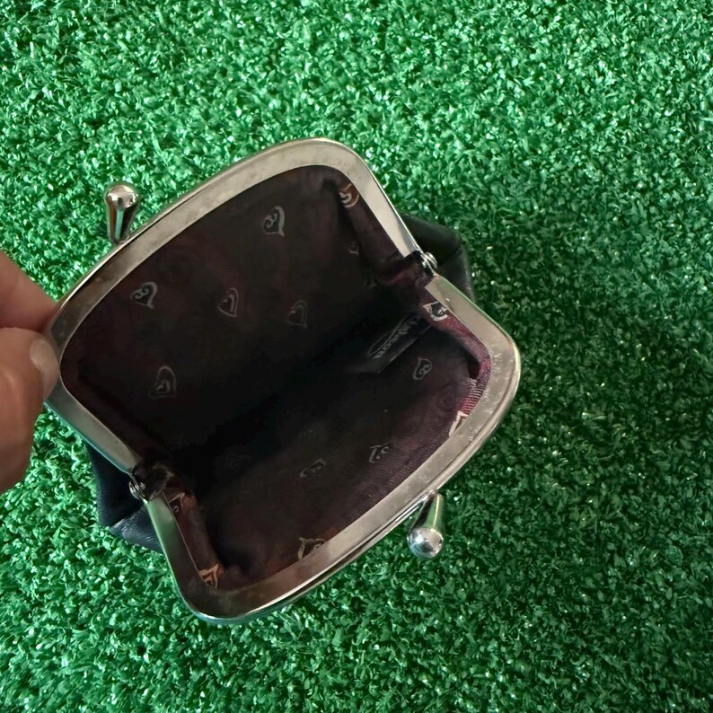 May include: A small black coin purse with a silver-toned metal kiss-lock frame. The interior is lined with dark brown fabric featuring a repeating pattern of small, light-colored heart-shaped icons and a visible brand tag inside the compartment.