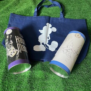 May include: Two blue and white metal tins with images of Mickey Mouse and characters from the Haunted Mansion attraction. The tins are sitting on top of a blue tote bag with a white silhouette of Mickey Mouse.