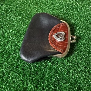 May include: A small coin purse featuring a smooth black leather body and a brown crocodile-textured leather flap. The flap is accented with a braided leather border and a silver-toned metal heart-shaped emblem. It has a silver-toned metal frame.