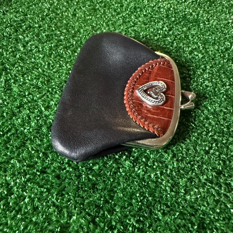 May include: A small coin purse featuring a smooth black leather body and a brown crocodile-textured leather flap. The flap is accented with a braided leather border and a silver-toned metal heart-shaped emblem. It has a silver-toned metal frame.