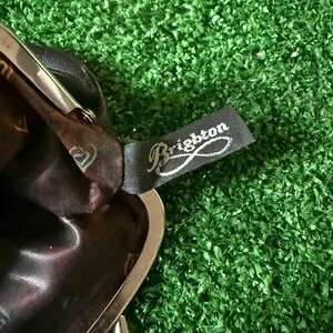 May include: A dark brown leather coin purse with a silver-toned metal frame and a kiss-lock closure. The interior is lined with a dark brown fabric featuring a repeating heart pattern. A black fabric tag with the white text Brighton is attached.