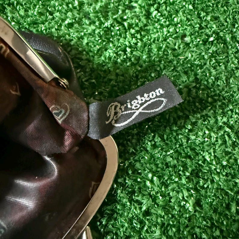 May include: A dark brown leather coin purse with a silver-toned metal frame and a kiss-lock closure. The interior is lined with a dark brown fabric featuring a repeating heart pattern. A black fabric tag with the white text Brighton is attached.
