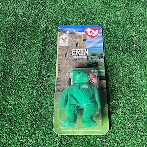 May include: A green plush bear toy named "Erin" with a shamrock on its chest, packaged in a clear plastic case. The packaging features a castle illustration and the text "Collect All 4!". The bear is on a green background.