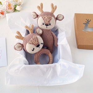 K&ouml;nnte beinhalten: Eine Geschenkbox gef&uuml;llt mit wei&szlig;em Seidenpapier enth&auml;lt zwei geh&auml;kelte Hirschspielzeuge. Ein Hirsch ist gr&ouml;&szlig;er und hat einen Holzring befestigt. Die Box ist braun mit einer ausgeschnittenen Hirschform. Eine Karte mit dem Text "Hello Baby" befindet sich im Hintergrund.