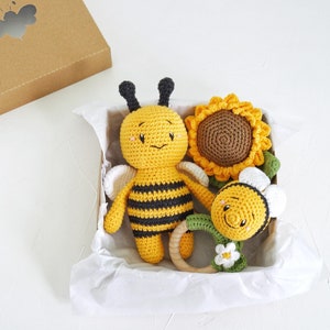 May include: A set of crocheted baby toys in a white gift box. The set includes a yellow and black bee, a yellow sunflower, and a yellow and white flower with a wooden teething ring.