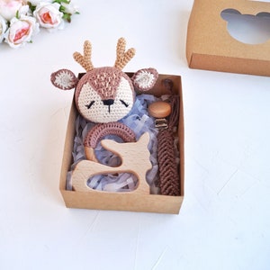 May include: A brown and beige crocheted deer rattle, a wooden teether shaped like a deer, and a brown crocheted teether with a wooden clip, all packaged in a brown box with a clear window.