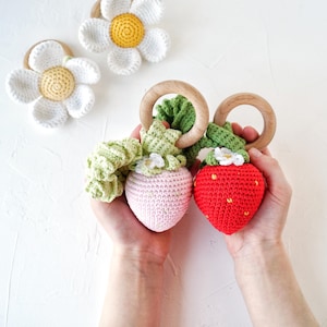 Peut inclure: Deux jouets en forme de fraise en crochet avec des anneaux en bois attachés. Une fraise est rose et l'autre est rouge.