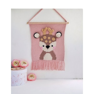 May include: A pink crocheted wall hanging with a brown deer face in the center. The deer has brown antlers, brown spots, and a white nose. The hanging has a fringe at the bottom.