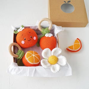 May include: A collection of orange crochet baby toys, including rattles and a flower, presented in a cardboard box. The toys feature wooden rings and white accents. The box has a die-cut orange shape on the lid.