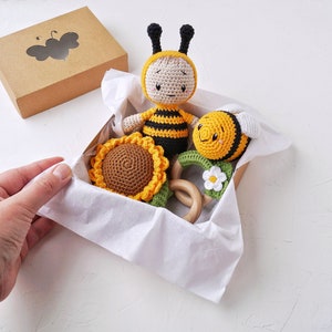 May include: A gift box filled with crocheted bee toys. The box is lined with white tissue paper and contains a yellow and black bee, a smaller yellow and black bee, a sunflower, and a wooden teething ring with a white flower.