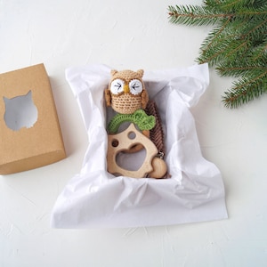 May include: A brown and green crocheted owl toy with a wooden teething ring and a brown and white knitted scarf in a white gift box.