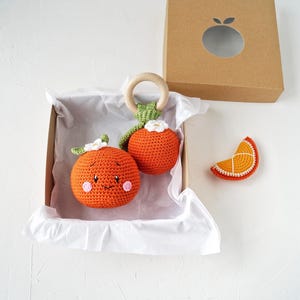 May include: A collection of orange crochet baby toys, including a rattle with a wooden ring and a slice of orange. The toys are presented in a cardboard box with an orange-shaped cut-out. The toys are orange, green, white, and brown.