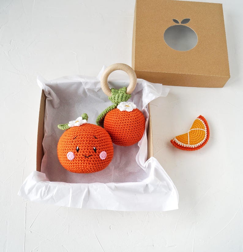 May include: A collection of orange crochet baby toys, including a rattle with a wooden ring and a slice of orange. The toys are presented in a cardboard box with an orange-shaped cut-out. The toys are orange, green, white, and brown.