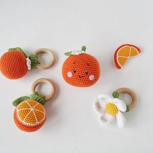 May include: A selection of handmade crochet baby toys shaped like oranges and a flower. The toys are orange, white, and green, with wooden rings. One orange has a smiling face and pink cheeks. The toys are designed for babies.