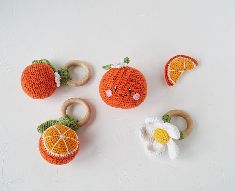 May include: A selection of handmade crochet baby toys shaped like oranges and a flower. The toys are orange, white, and green, with wooden rings. One orange has a smiling face and pink cheeks. The toys are designed for babies.