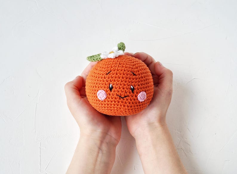May include: A crocheted orange fruit with a smiling face, pink cheeks, and a green and white leaf and flower detail. The orange is held in two hands against a white background.